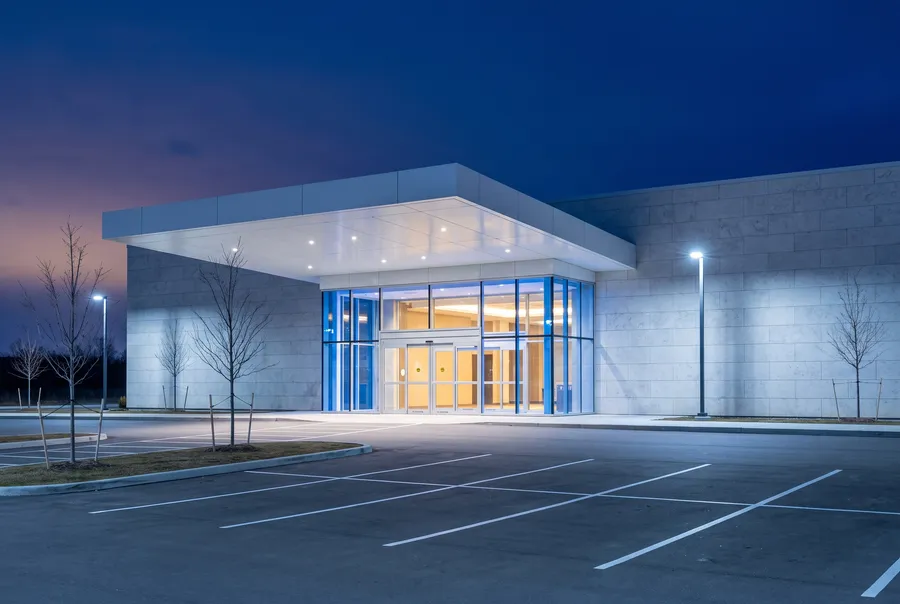 A modern single-storey casino entrance at blue hour: a flat white canopy over glass doors with warm light inside, pale stone walls, an empty parking lot with fresh white lines